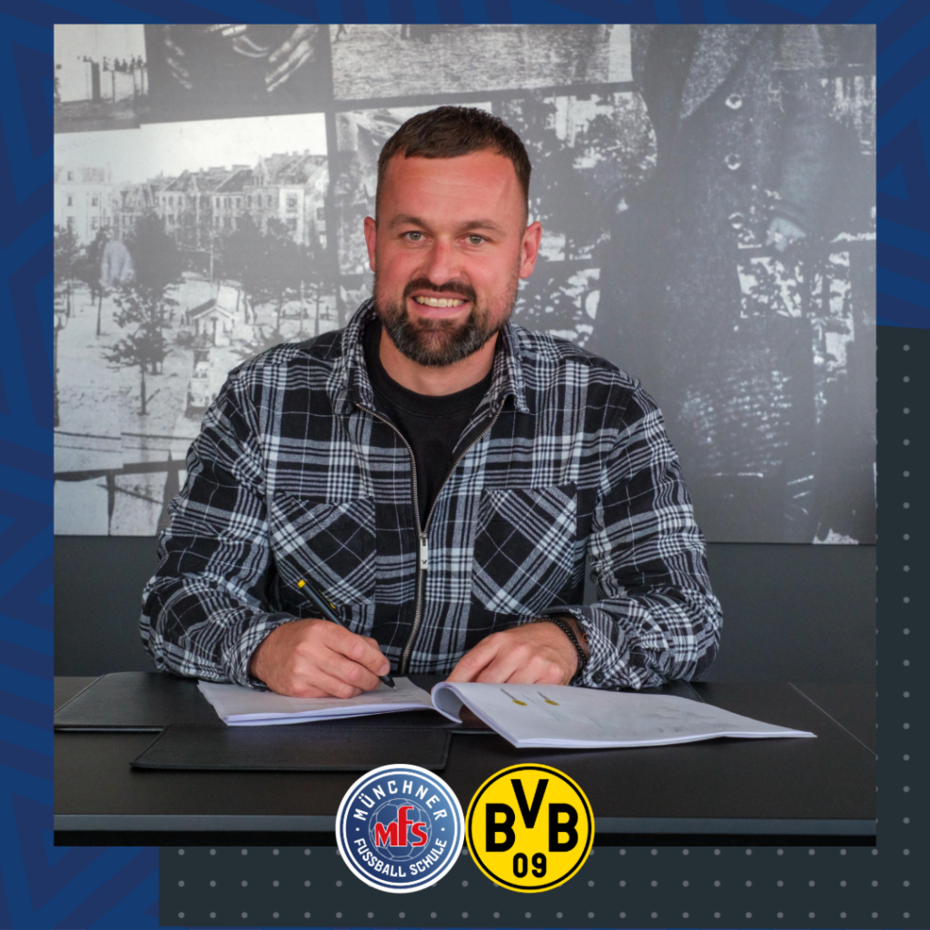 Ein Mann mit Bart und einem schwarz-weiß karierten Hemd lächelt, während er an einem Tisch ein Dokument unterschreibt.